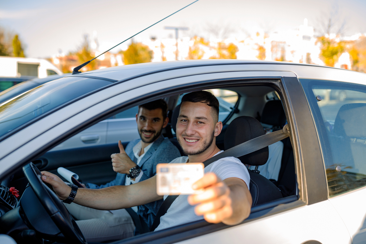 A young Caucasian male driving instructor holding thumbs-up and a young Caucasian man holding a new drivers license are sitting in a car and looking at the camera with smiles on their faces.